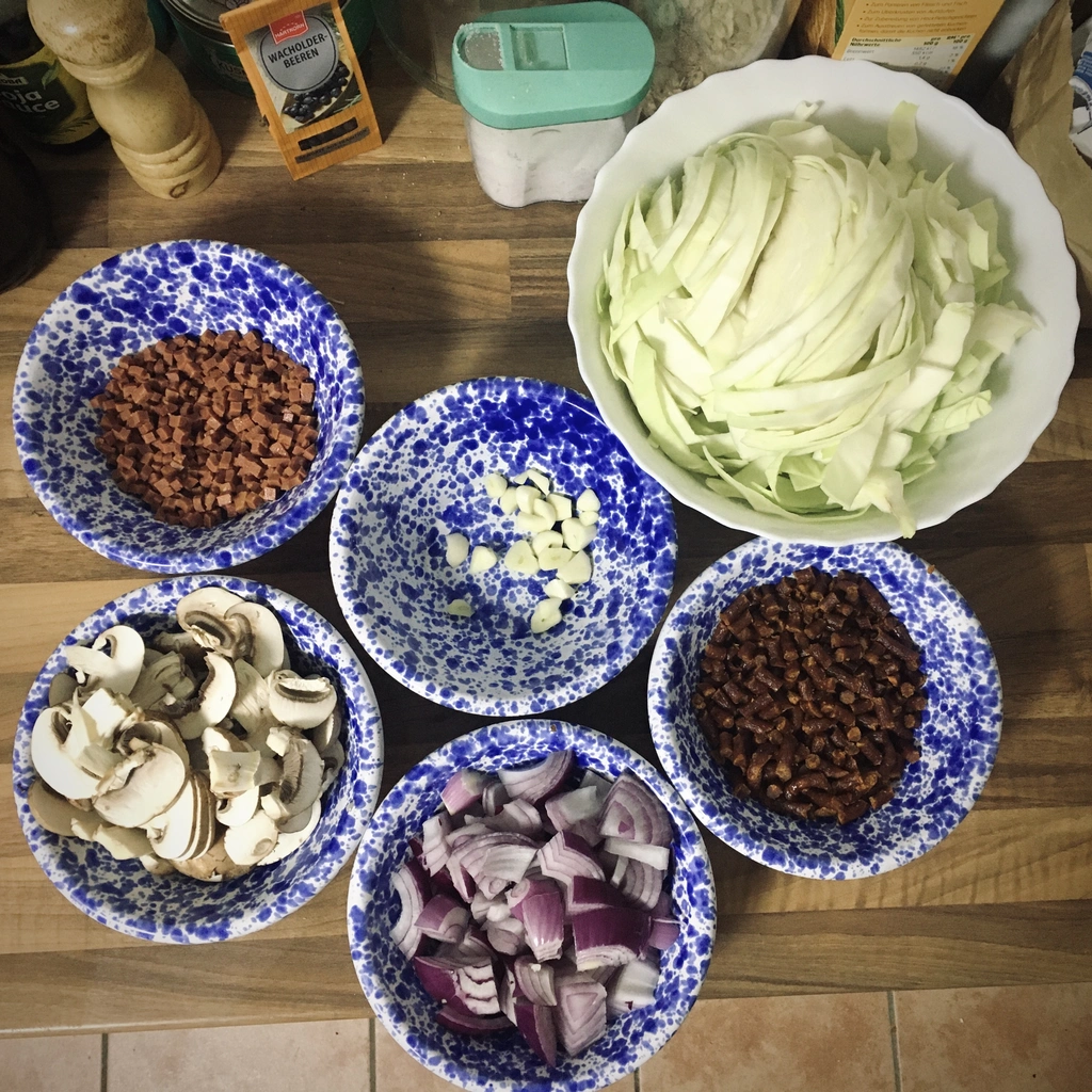 Mehrere Schalen mit klein geschnittenen Gemüse und veganen Speck/Kabanossi, sowie eine große Schüssel mit Weißkohl, welches in Streifen geschnitten wurde.