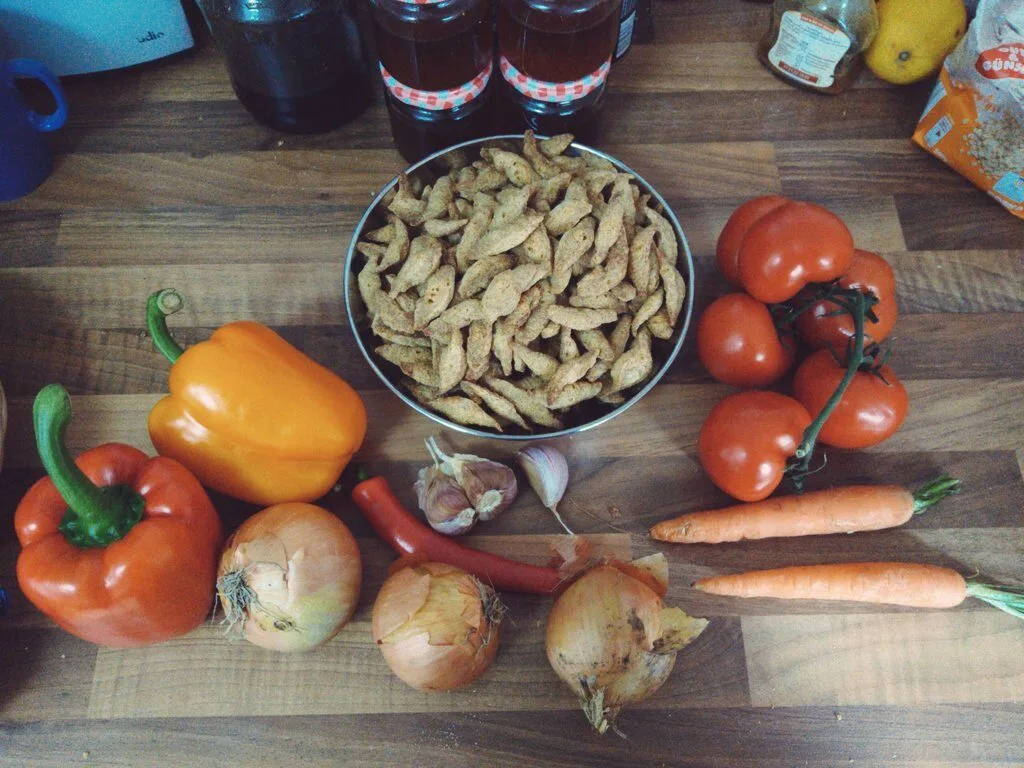 Zutaten für das Gulasch. Soja Schnetzel in einer metalischen Schüssel, Paprika in Rot und Orange, drei Zwiebel, Chilischote, Knoblauch, Tomaten und Möhren