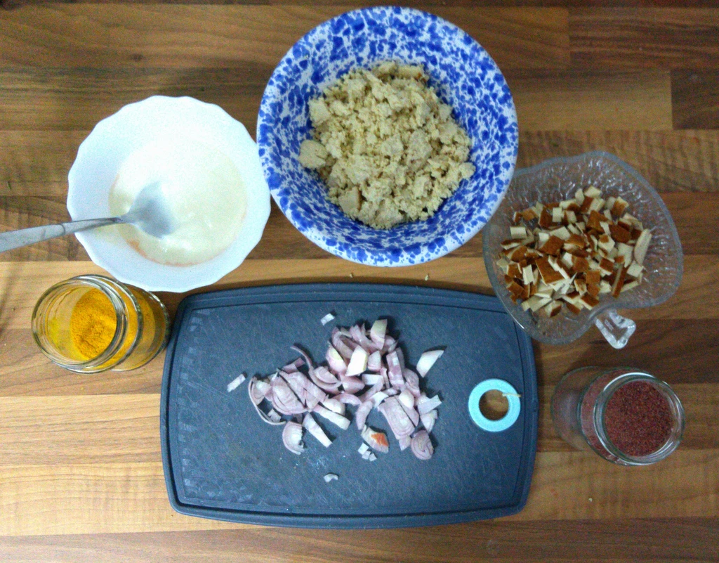 Joghurt, Tofu und Räucherkruste jeweils in einer Schale. Links ist ein Glas mit Kurkuma und eines mit Kala Namak. Auf einem Brettchen ist eine Zwiebel klein geschnitten.